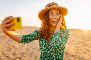 Mutlu turist, deniz manzarasında selfie çekiyor. Güzel bir kadın sahilde gün batımının tadını çıkarıyor. Telefonla, blogla. Hafta sonu, günlük tutma kavramı.