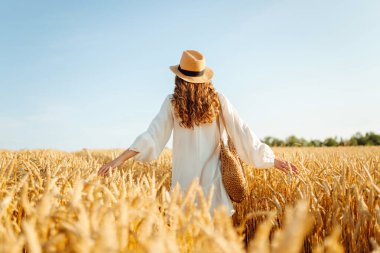 Beyaz elbiseli ve şapkalı genç bir kadının altın buğday tarlasında günbatımının keyfini çıkarırken görüntüsü. Güzel bir kadın kendini özgür hisseder ve tarımdan geçer. Çiftçilik ve bahçecilik kavramı.