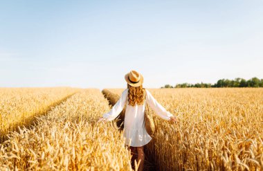 Beyaz elbiseli ve şapkalı genç bir kadının altın buğday tarlasında günbatımının keyfini çıkarırken görüntüsü. Güzel bir kadın kendini özgür hisseder ve tarımdan geçer. Çiftçilik ve bahçecilik kavramı.