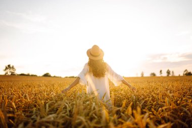 Beyaz elbiseli ve şapkalı genç bir kadının altın buğday tarlasında günbatımının keyfini çıkarırken görüntüsü. Güzel bir kadın kendini özgür hisseder ve tarımdan geçer. Çiftçilik ve bahçecilik kavramı.