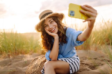 Şapkalı, modern elbiseli ve telefonlu neşeli kadın sahilde günbatımının tadını çıkarıyor. Güzel bir turist doğada selfie çeker ve blog yazar. Keyif ve eğlence konsepti.
