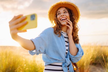 Şapkalı, modern elbiseli ve telefonlu neşeli kadın sahilde günbatımının tadını çıkarıyor. Güzel bir turist doğada selfie çeker ve blog yazar. Keyif ve eğlence konsepti.