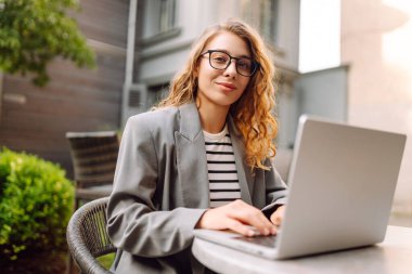 Dürbünlü güzel bir kadın dışarıda, masada çalışıyor. Kafede dizüstü bilgisayarı olan serbest çalışan bir kadın havanın tadını çıkarıyor. Teknoloji konsepti, uzaktan çalışma.