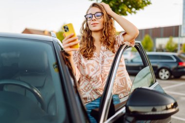 Park yerinde arabasının yanında gözlüklü ve telefonu olan genç bir kadın. Telefonlu güzel bir kadın, gün batımında blog yazıyor. Dinlenme kavramı, rahatlama..