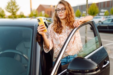 Park yerinde arabasının yanında gözlüklü ve telefonu olan genç bir kadın. Telefonlu güzel bir kadın, gün batımında blog yazıyor. Dinlenme kavramı, rahatlama..