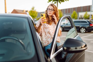 Park yerinde arabasının yanında gözlüklü ve telefonu olan genç bir kadın. Telefonlu güzel bir kadın, gün batımında blog yazıyor. Dinlenme kavramı, rahatlama..