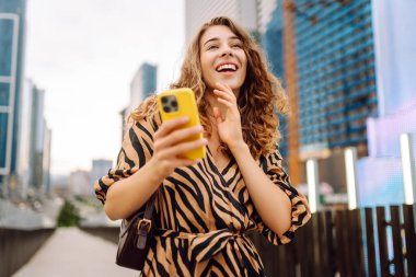 Şık giyinmiş, şehirli bir çevrede telefonla yürüyen, yüksek binalara bakan genç bir kadının portresi. Güzel bir turist havanın tadını çıkarıyor ve dışarıda blog yazıyor. Turizm konsepti.