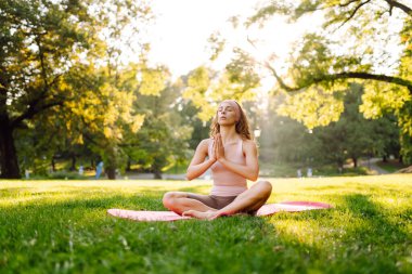 Güzel bir kadın parkta pembe bir paspasın üzerinde esneme egzersizleri yapıyor. Gün batımında meditasyon yapan genç bir kadın. Dinlenme ve rahatlama kavramı.