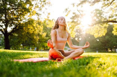 Güzel bir kadın parkta pembe bir paspasın üzerinde esneme egzersizleri yapıyor. Gün batımında meditasyon yapan genç bir kadın. Dinlenme ve rahatlama kavramı.
