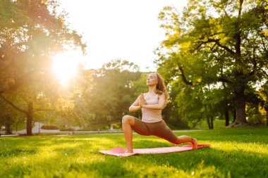 Güzel bir kadın parkta pembe bir paspasın üzerinde esneme egzersizleri yapıyor. Gün batımında meditasyon yapan genç bir kadın. Dinlenme ve rahatlama kavramı.