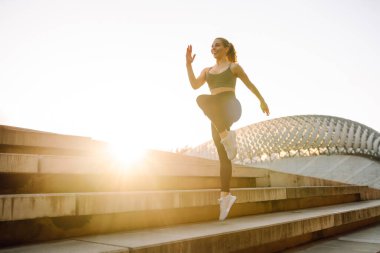 Spor kıyafetli genç bir kadın gün batımında merdivenlerde egzersiz yapıyor. Atletik kadın dışarıda zıplama hareketleri yapıyor. Spor konsepti. Etkin yaşam biçimi.