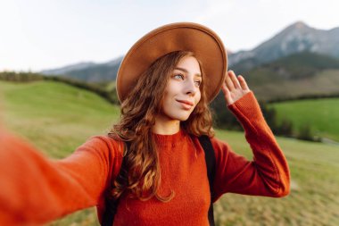 Yeşil dağ tepesinde selfie çeken şapkalı kadın portresi. Genç bayan gezgin blog yazıyor ve günbatımının tadını çıkarıyor. Macera, blog ve özgürlük kavramı.