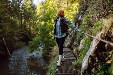 Genç bir kadın sık bir ormanda nehir boyunca metal bir patikada yürüyor. Güzel bayan gezgin gün batımının ve dağ manzarasının tadını çıkarır. Yürüyüş, eğlence konsepti.