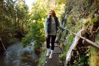 Genç bir kadın sık bir ormanda nehir boyunca metal bir patikada yürüyor. Güzel bayan gezgin gün batımının ve dağ manzarasının tadını çıkarır. Yürüyüş, eğlence konsepti.