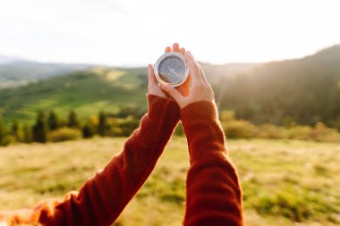 Şapkalı ve elinde pusula olan güzel bir kadın çarpıcı manzaralı yeşil bir tepede duruyor. Genç gezgin gün batımının tadını çıkarıyor ve yol tarifi arıyor. Seyahat kavramı.