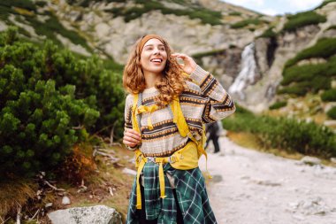 Arka planda bir dağ şelalesi ile yürüyüş yolunda yürüyen renkli bir sırt çantasıyla genç bir bayan gezgin. Doğanın tadını çıkaran ve özgürlüğü hisseden güzel bir kadın. Yürüyüş kavramı.