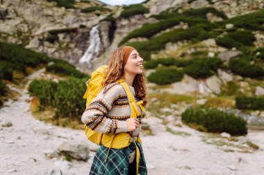Arka planda bir dağ şelalesi ile yürüyüş yolunda yürüyen renkli bir sırt çantasıyla genç bir bayan gezgin. Doğanın tadını çıkaran ve özgürlüğü hisseden güzel bir kadın. Yürüyüş kavramı.