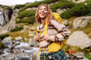 Arka planda bir dağ şelalesi ile yürüyüş yolunda yürüyen renkli bir sırt çantasıyla genç bir bayan gezgin. Doğanın tadını çıkaran ve özgürlüğü hisseden güzel bir kadın. Yürüyüş kavramı.