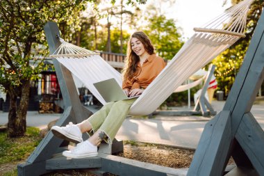 Şık giyinmiş, dizüstü bilgisayarı hamakta dinlenen genç bir kadın. Parkta çalışan güzel bir serbest çalışan kadın. Uzak çalışma ve boş zaman kavramı.