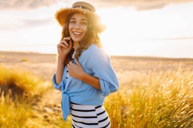 Genç bir kadın sahilde günbatımının tadını çıkarıyor. Şapkalı ve çantalı şık bir kadın deniz kenarında yürüyor. Yürüyüş, güzellik ve tarz kavramı.