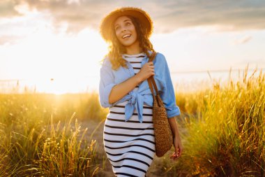 Genç bir kadın sahilde günbatımının tadını çıkarıyor. Şapkalı ve çantalı şık bir kadın deniz kenarında yürüyor. Yürüyüş, güzellik ve tarz kavramı.