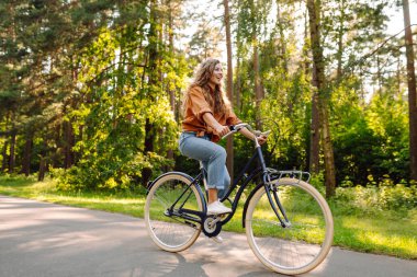 Kıvırcık saçlı neşeli bir kadın güneşli bir parkta bisiklet sürüyor. Genç bir kadın dışarıda eğleniyor ve eğleniyor. Etkin yaşam biçimi.