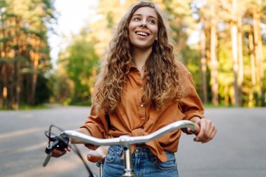 Kıvırcık saçlı neşeli bir kadın güneşli bir parkta bisiklet sürüyor. Genç bir kadın dışarıda eğleniyor ve eğleniyor. Etkin yaşam biçimi.