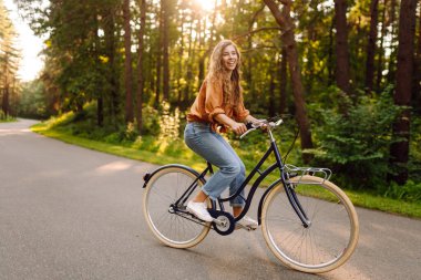 Kıvırcık saçlı neşeli bir kadın güneşli bir parkta bisiklet sürüyor. Genç bir kadın dışarıda eğleniyor ve eğleniyor. Etkin yaşam biçimi.