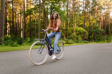 Kıvırcık saçlı neşeli bir kadın güneşli bir parkta bisiklet sürüyor. Genç bir kadın dışarıda eğleniyor ve eğleniyor. Etkin yaşam biçimi.