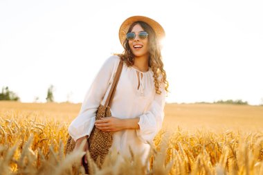 Şapkalı ve güneş gözlüklü mutlu bir kadının portresi altın tarlada eğleniyor. Turist buğdayda gün batımının tadını çıkarıyor, özgürlüğü hissediyor. Tarz ve güzellik konsepti, rahatlama.
