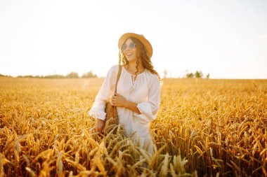 Şapkalı ve güneş gözlüklü mutlu bir kadının portresi altın tarlada eğleniyor. Turist buğdayda gün batımının tadını çıkarıyor, özgürlüğü hissediyor. Tarz ve güzellik konsepti, rahatlama.