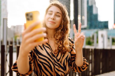 Selfie zamanı! Telefonu olan güzel bir kadın, gökdelenlere tepeden bakan gezinti güvertesinde eğleniyor. Genç bir blogcu gün batımını dışarıda izlemeyi sever. Turizm konsepti.