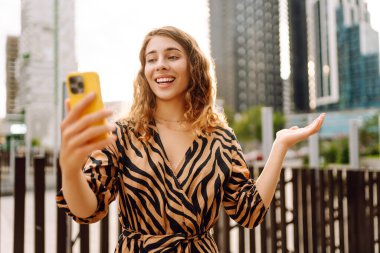 Selfie zamanı! Telefonu olan güzel bir kadın, gökdelenlere tepeden bakan gezinti güvertesinde eğleniyor. Genç bir blogcu gün batımını dışarıda izlemeyi sever. Turizm konsepti.