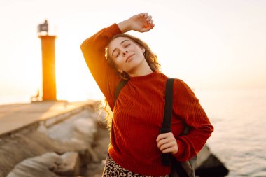 Sırt çantalı mutlu bir kadın deniz fenerine tepeden bakan bir iskelede özgür hisseder. Genç bir turist denizde günbatımının tadını çıkarıyor. Keyif konsepti, rahatlama.