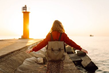Sırt çantalı mutlu bir kadın deniz fenerine tepeden bakan bir iskelede özgür hisseder. Genç bir turist denizde günbatımının tadını çıkarıyor. Keyif konsepti, rahatlama.