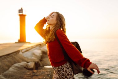 Sırt çantalı mutlu bir kadın deniz fenerine tepeden bakan bir iskelede özgür hisseder. Genç bir turist denizde günbatımının tadını çıkarıyor. Keyif konsepti, rahatlama.