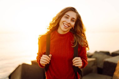 Sırt çantalı mutlu bir kadın deniz fenerine tepeden bakan bir iskelede özgür hisseder. Genç bir turist denizde günbatımının tadını çıkarıyor. Keyif konsepti, rahatlama.