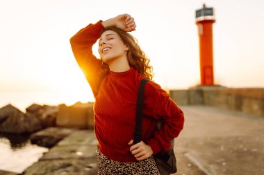 Sırt çantalı mutlu bir kadın deniz fenerine tepeden bakan bir iskelede özgür hisseder. Genç bir turist denizde günbatımının tadını çıkarıyor. Keyif konsepti, rahatlama.