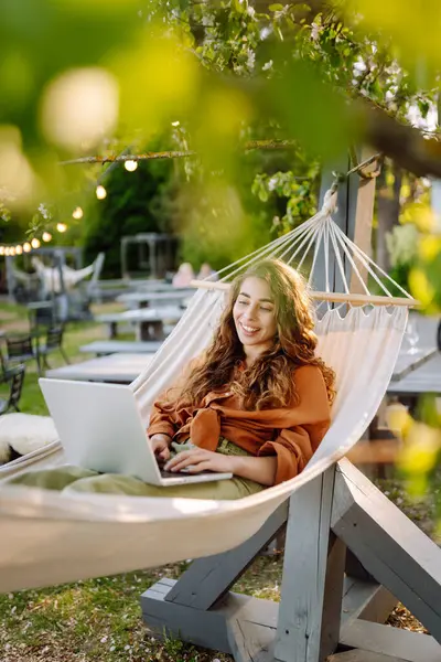 Şık giyinmiş, dizüstü bilgisayarı hamakta dinlenen genç bir kadın. Parkta çalışan güzel bir serbest çalışan kadın. Uzak çalışma ve boş zaman kavramı.
