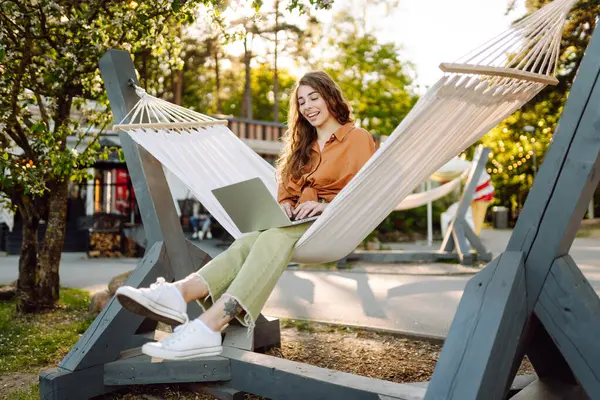 Şık giyinmiş, dizüstü bilgisayarı hamakta dinlenen genç bir kadın. Parkta çalışan güzel bir serbest çalışan kadın. Uzak çalışma ve boş zaman kavramı.
