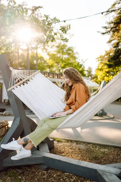 Şık giyinmiş, dizüstü bilgisayarı hamakta dinlenen genç bir kadın. Parkta çalışan güzel bir serbest çalışan kadın. Uzak çalışma ve boş zaman kavramı.