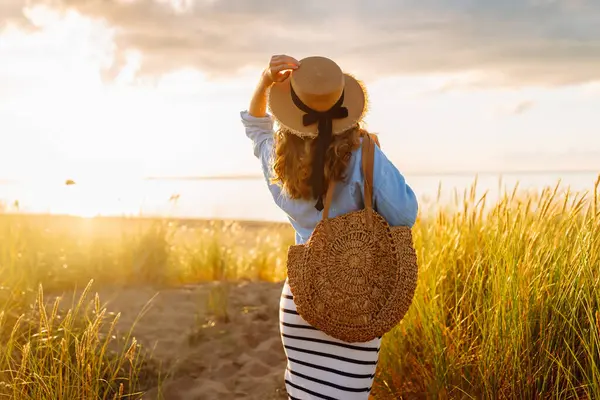 Genç bir kadın sahilde günbatımının tadını çıkarıyor. Şapkalı ve çantalı şık bir kadın deniz kenarında yürüyor. Yürüyüş, güzellik ve tarz kavramı.