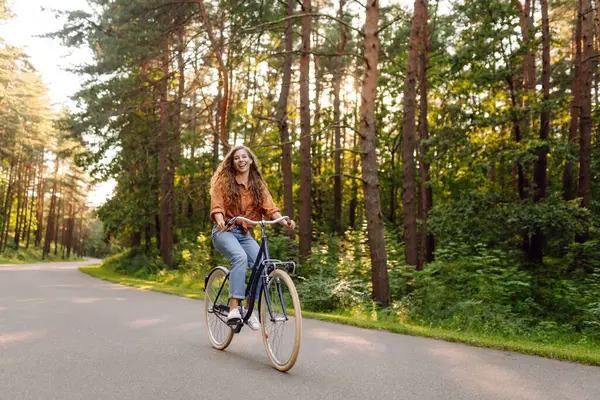 Kıvırcık saçlı neşeli bir kadın güneşli bir parkta bisiklet sürüyor. Genç bir kadın dışarıda eğleniyor ve eğleniyor. Etkin yaşam biçimi.
