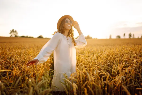 Şapkalı ve güneş gözlüklü mutlu bir kadının portresi altın tarlada eğleniyor. Turist buğdayda gün batımının tadını çıkarıyor, özgürlüğü hissediyor. Tarz ve güzellik konsepti, rahatlama.