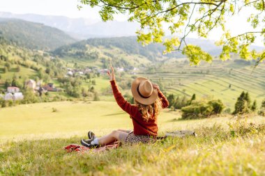 Çimenli yamaçta oturan genç bir kadının arka görüntüsü. Şapkalı gezgin dağ manzarasının ve doğada gün batımının tadını çıkarıyor. Özgürlük kavramı. Etkin yaşam biçimi.