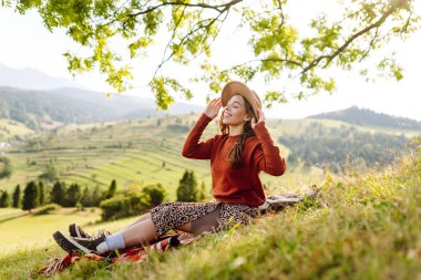 Çimenli yamaçta oturan genç bir kadının arka görüntüsü. Şapkalı gezgin dağ manzarasının ve doğada gün batımının tadını çıkarıyor. Özgürlük kavramı. Etkin yaşam biçimi.