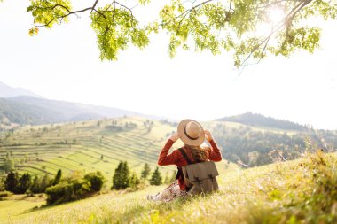 Çimenli yamaçta oturan genç bir kadının arka görüntüsü. Şapkalı gezgin dağ manzarasının ve doğada gün batımının tadını çıkarıyor. Özgürlük kavramı. Etkin yaşam biçimi.
