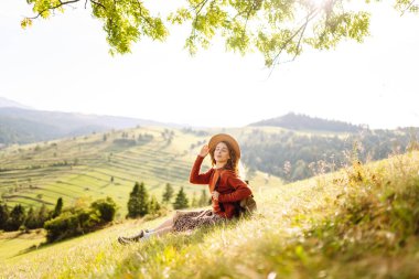 Çimenli yamaçta oturan genç bir kadının arka görüntüsü. Şapkalı gezgin dağ manzarasının ve doğada gün batımının tadını çıkarıyor. Özgürlük kavramı. Etkin yaşam biçimi.