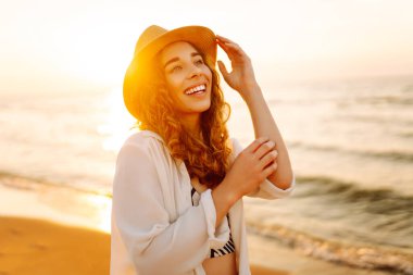 Sahilde günbatımının tadını çıkaran şapkalı güzel bir kadının portresi. Genç kadın özgürlüğü hissediyor, doğada rahatlıyor. Keyif ve rahatlama kavramı.