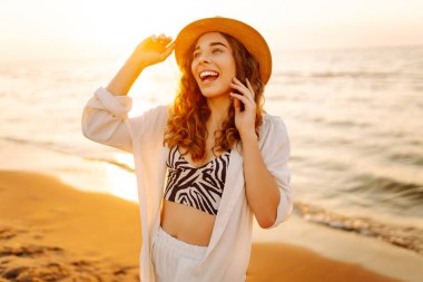 Sahilde günbatımının tadını çıkaran şapkalı güzel bir kadının portresi. Genç kadın özgürlüğü hissediyor, doğada rahatlıyor. Keyif ve rahatlama kavramı.